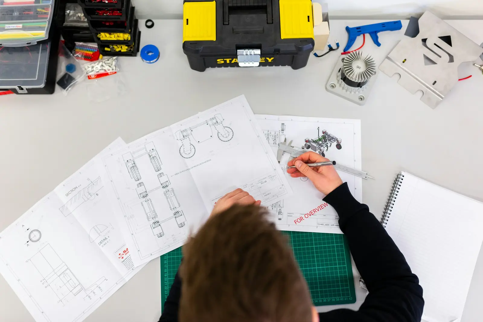 Vue de dessus d'une personne qui annote des plans techniques sur un plan de travail, à côté d'une caisse à outils